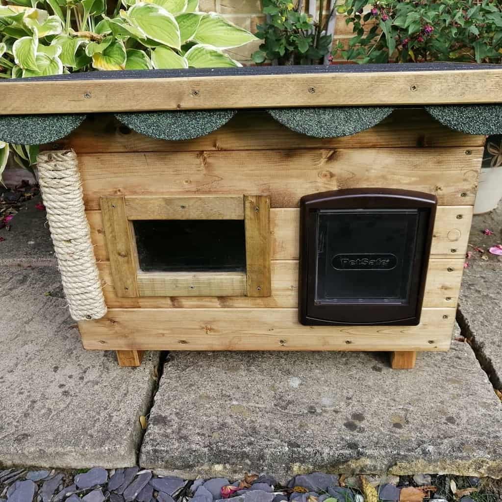 Wooden cat house with clear window, brown pet door, and rope scratching post on the side surrounded by garden plants