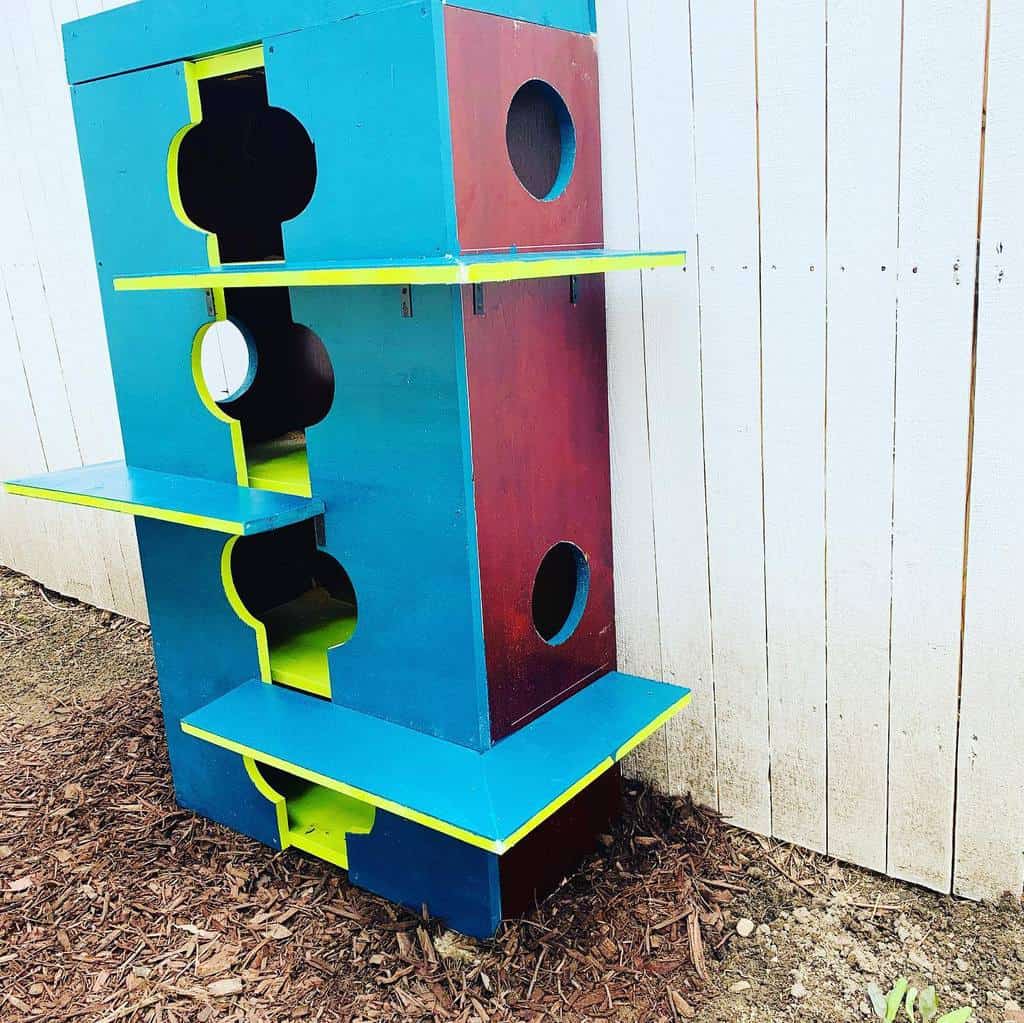 Colorful multi-level wooden cat tree with circular openings, positioned against a white fence