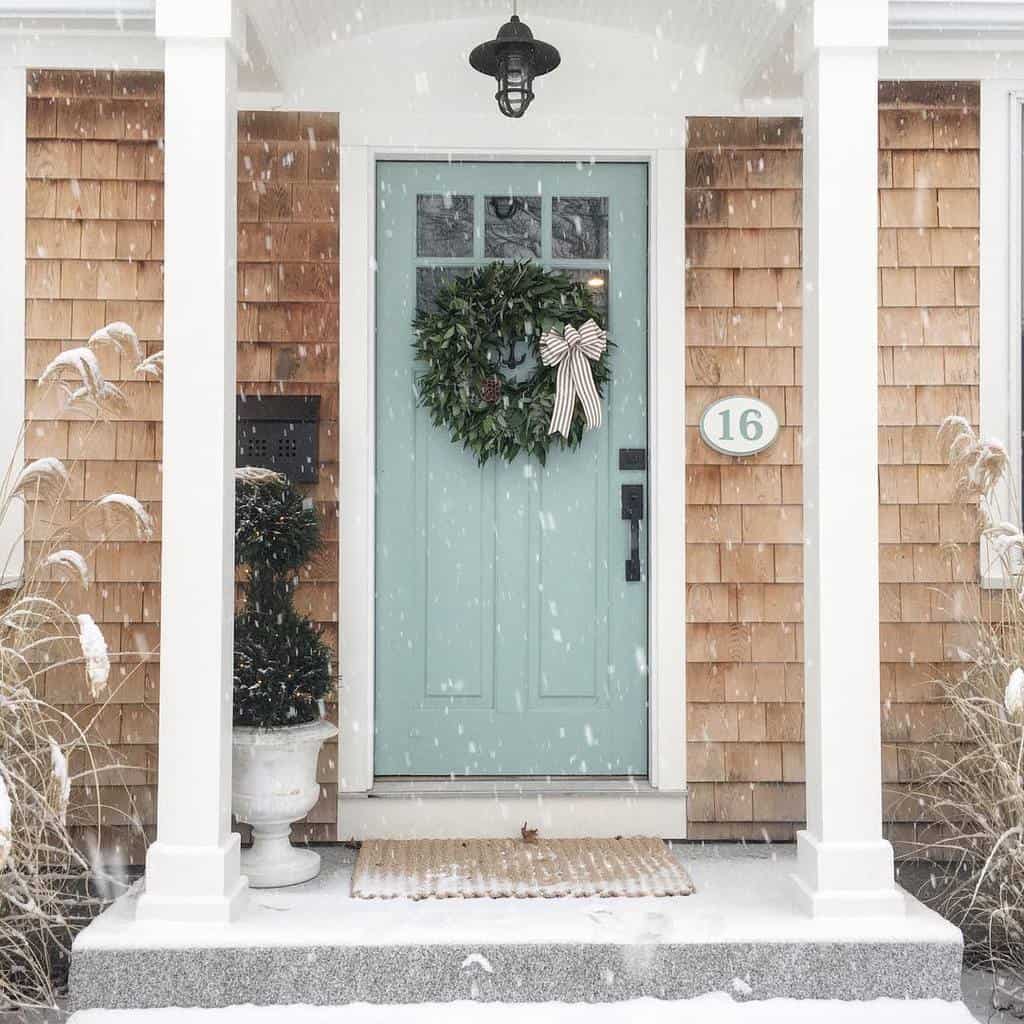 Snowy front porch with a light blue door, green wreath with a striped bow, and light wood siding