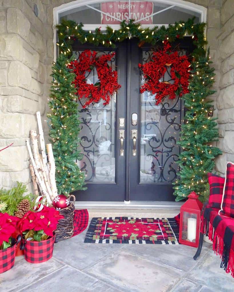 Festive porch with double doors, red wreaths, garland, poinsettias, a lantern, plaid decor, and a "Merry Christmas" sign