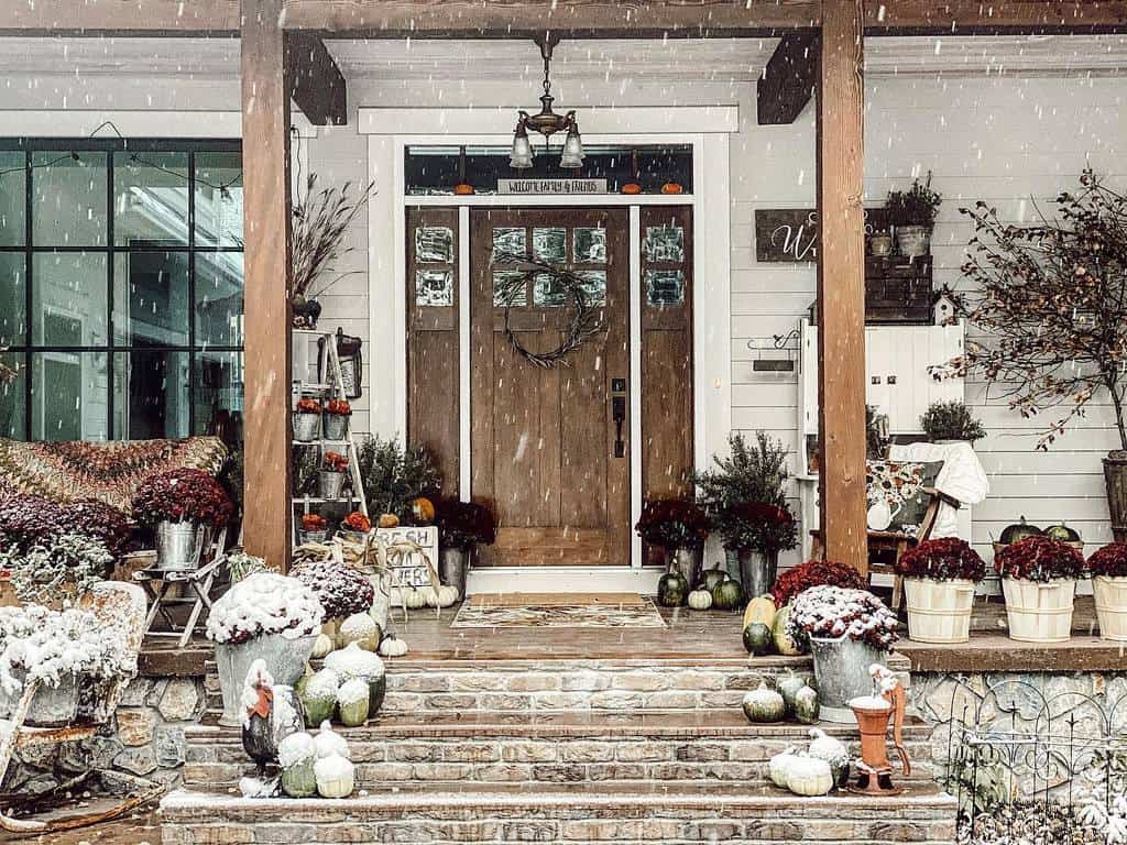 Cozy porch decorated for fall, with pumpkins, plants, and flowers under a light snowfall; wooden door and stone steps enhance the scene