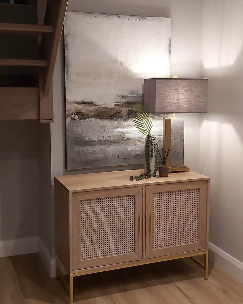 Modern entryway with a woven cane console, abstract artwork, a warm table lamp, and decorative greenery for a stylish, inviting look