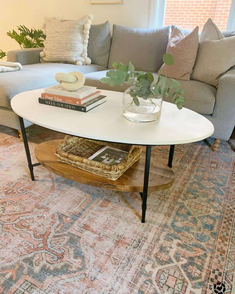 Modern oval coffee table with a white top, black metal legs, and a wooden lower shelf, styled with books, a vase, and a woven tray