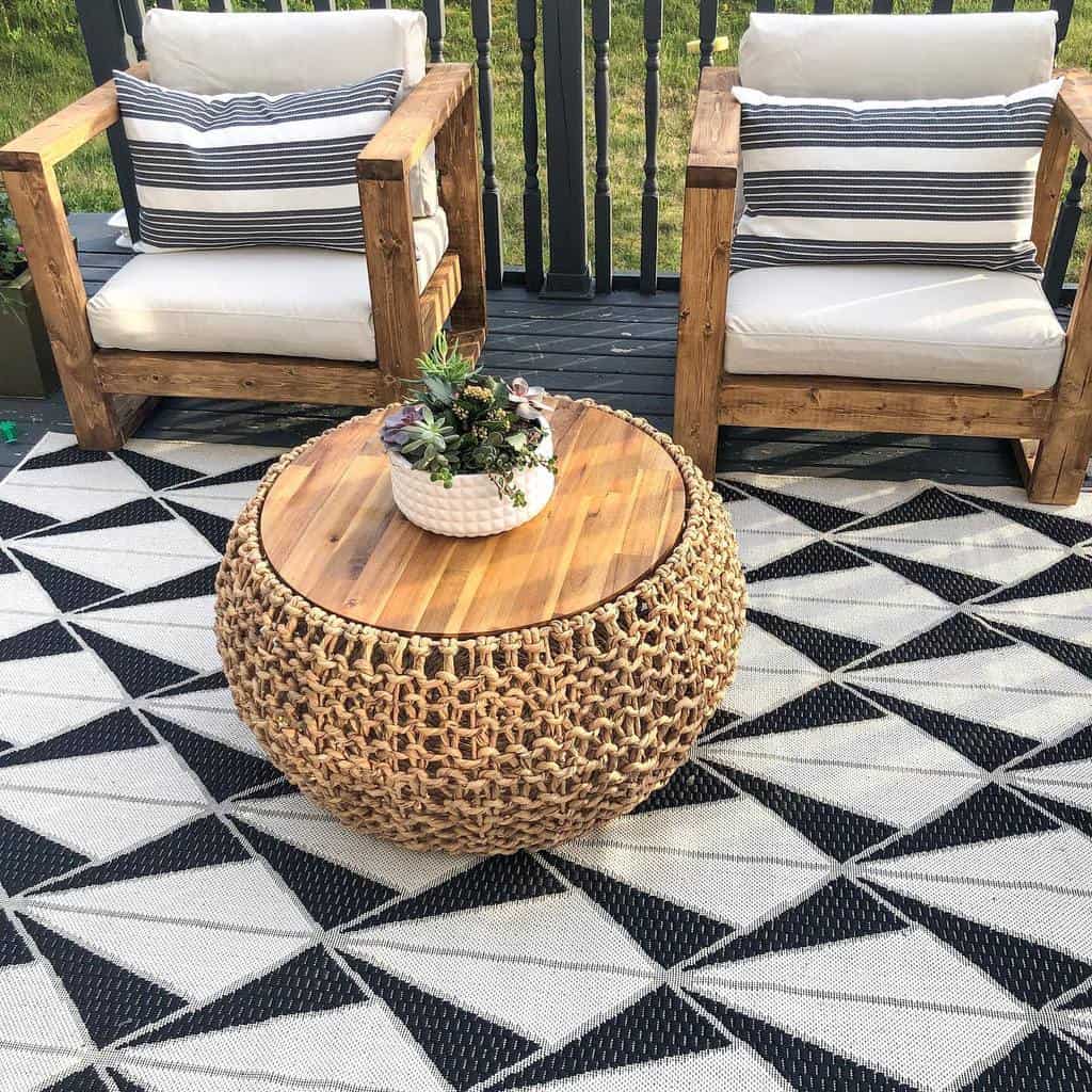 Outdoor seating area with two wooden armchairs, black and white striped cushions, and a woven round table with a plant on top; geometric rug