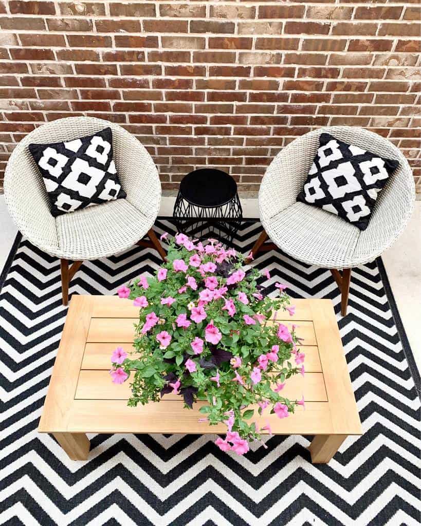 Patio with two white chairs and geometric cushions, black side table, wooden coffee table, vibrant pink flowers, and brick wall backdrop