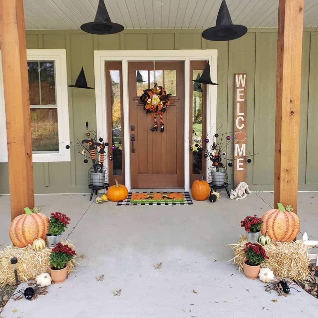 Front porch with Halloween decor: pumpkins, hay bales, witch hats, plants, and a "WELCOME" sign beside a door adorned with a wreath