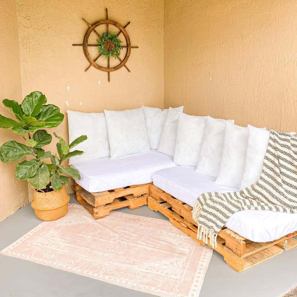 Cozy corner with a white-cushioned pallet sofa, plant in a woven basket, a striped throw, and a wooden ship wheel wall decor