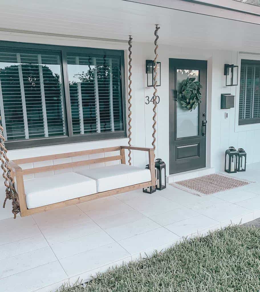 Front porch with a wooden swing, white cushions, gray door with wreath, lanterns on the ground, and neatly trimmed grass