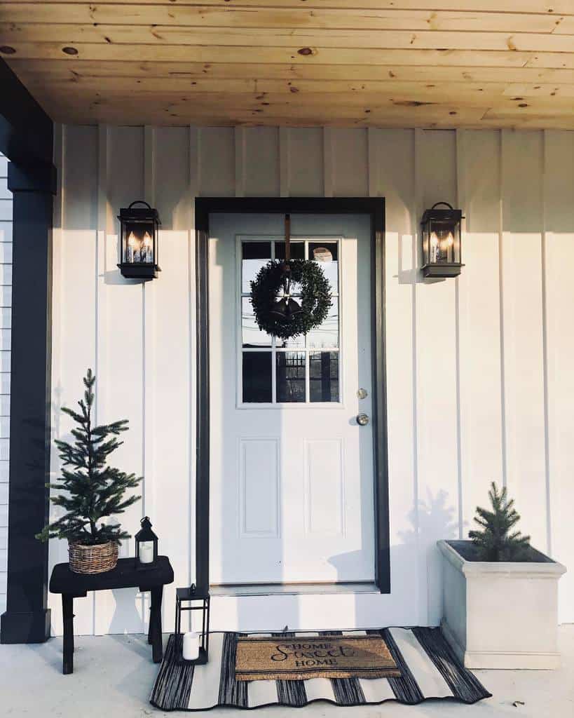 Front porch with a white door, wreath, two small evergreen trees, lanterns, and a "Home Sweet Home" doormat on a striped rug