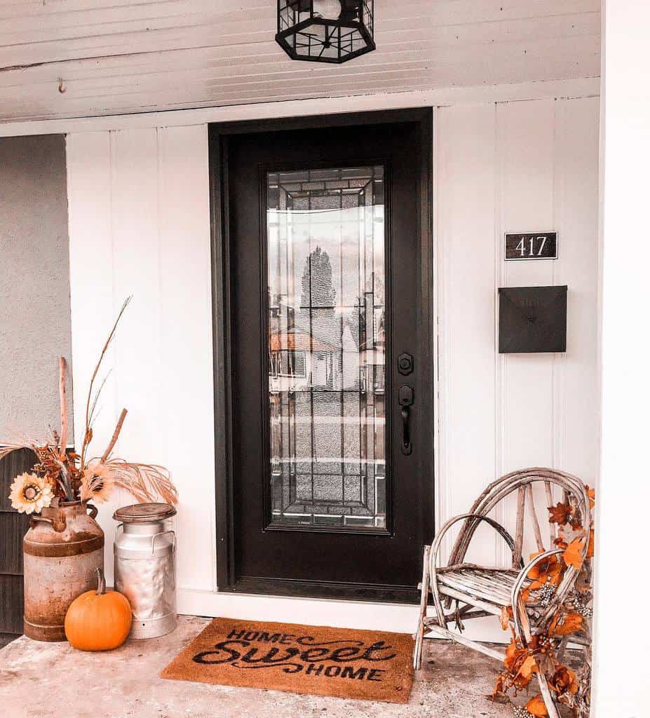 Front porch with black door, welcome mat reading "Home Sweet Home," rustic chair, and fall decor including pumpkin and dried flowers