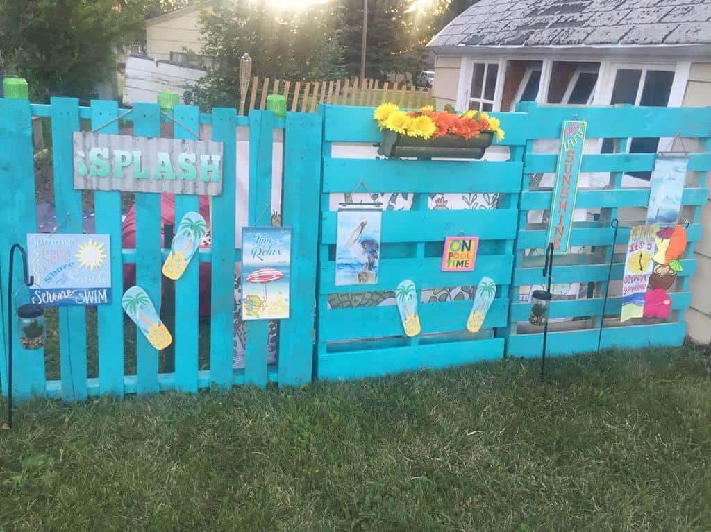 Bright turquoise DIY pallet fence decorated with summer-themed signs, flip-flops, and flowers, creating a fun backyard oasis