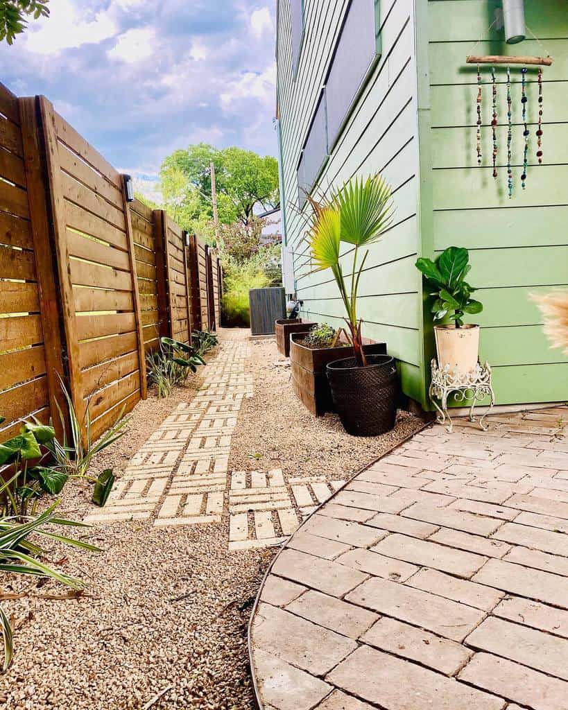 Side walkway with potted plants and a hanging decoration on a light green house, bordered by a wooden fence and paved with bricks