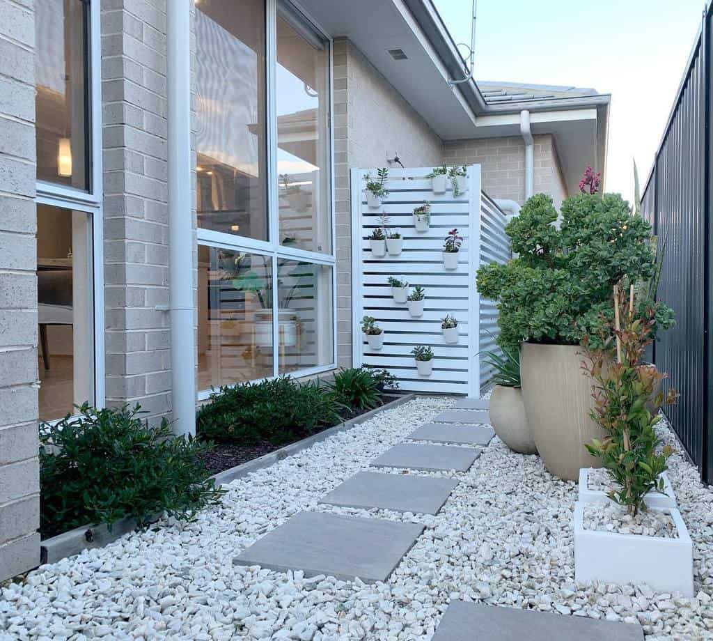 Modern narrow garden with stepping stones, white gravel, potted plants, and a wooden panel decorated with small planters