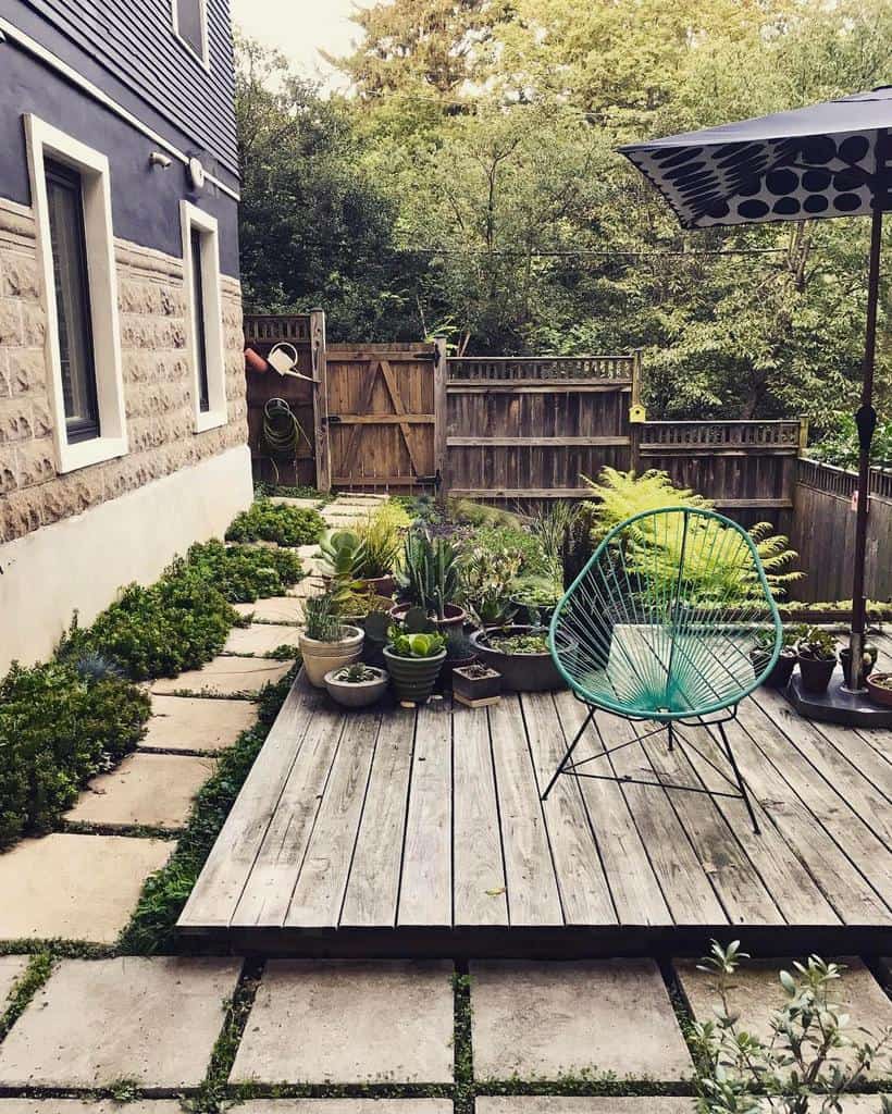 Side yard with a wooden deck, potted succulents, stone pathway, turquoise chair, and a black patio umbrella, enclosed by a wooden fence