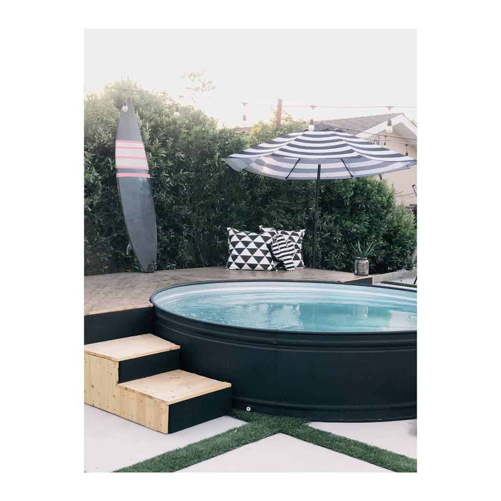 Backyard with a round above-ground pool, wooden steps, surfboard, two patterned pillows, and a striped umbrella