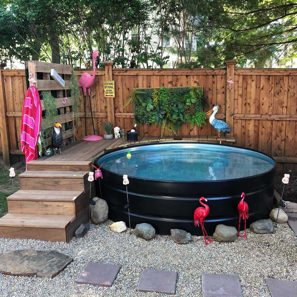 Backyard pool setup with wooden steps, pink flamingo decor, plants, and a wooden fence. Rocks and gravel surround the pool