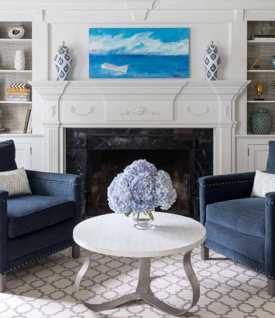 Round marble coffee table with curved metal legs, topped with a vase of blue hydrangeas, set between navy armchairs by a fireplace