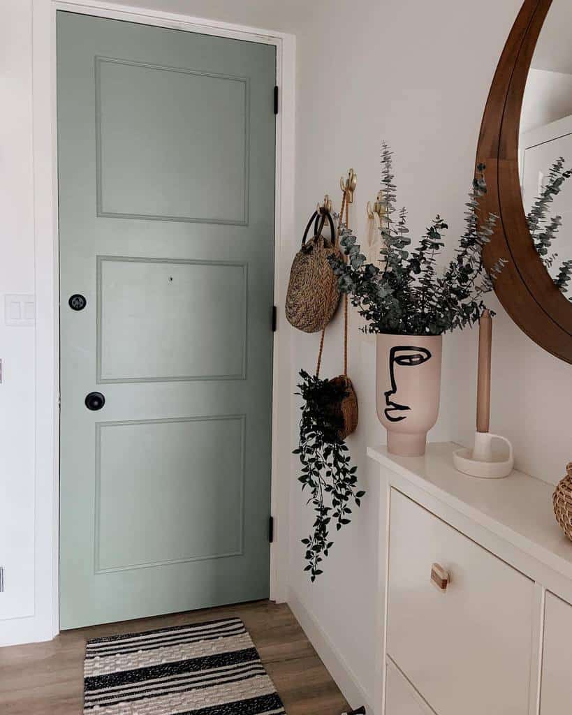 Chic entryway with a sage green door, minimalist storage, gold wall hooks, a modern face vase with greenery, and a striped rug