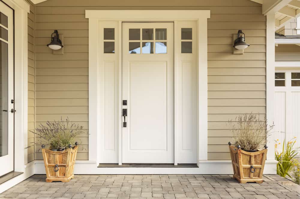 A white front door with glass panes, flanked by wooden planters and wall lanterns, under a porch with beige siding