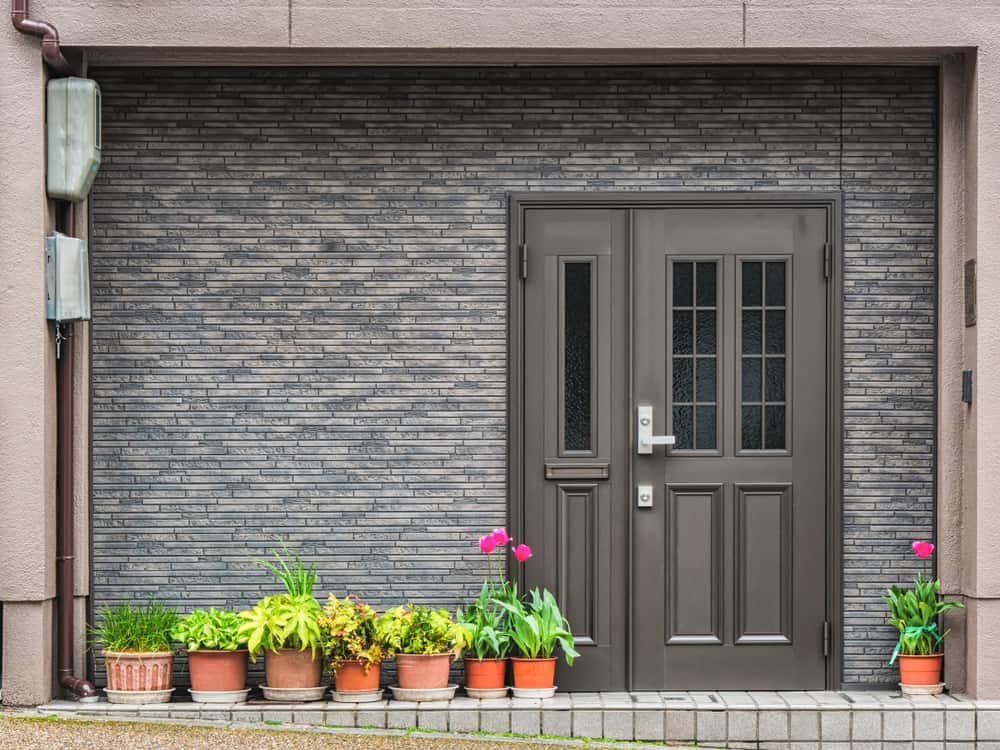 Gray brick wall with a dark brown door, adorned with potted plants and flowers on both sides of the entrance