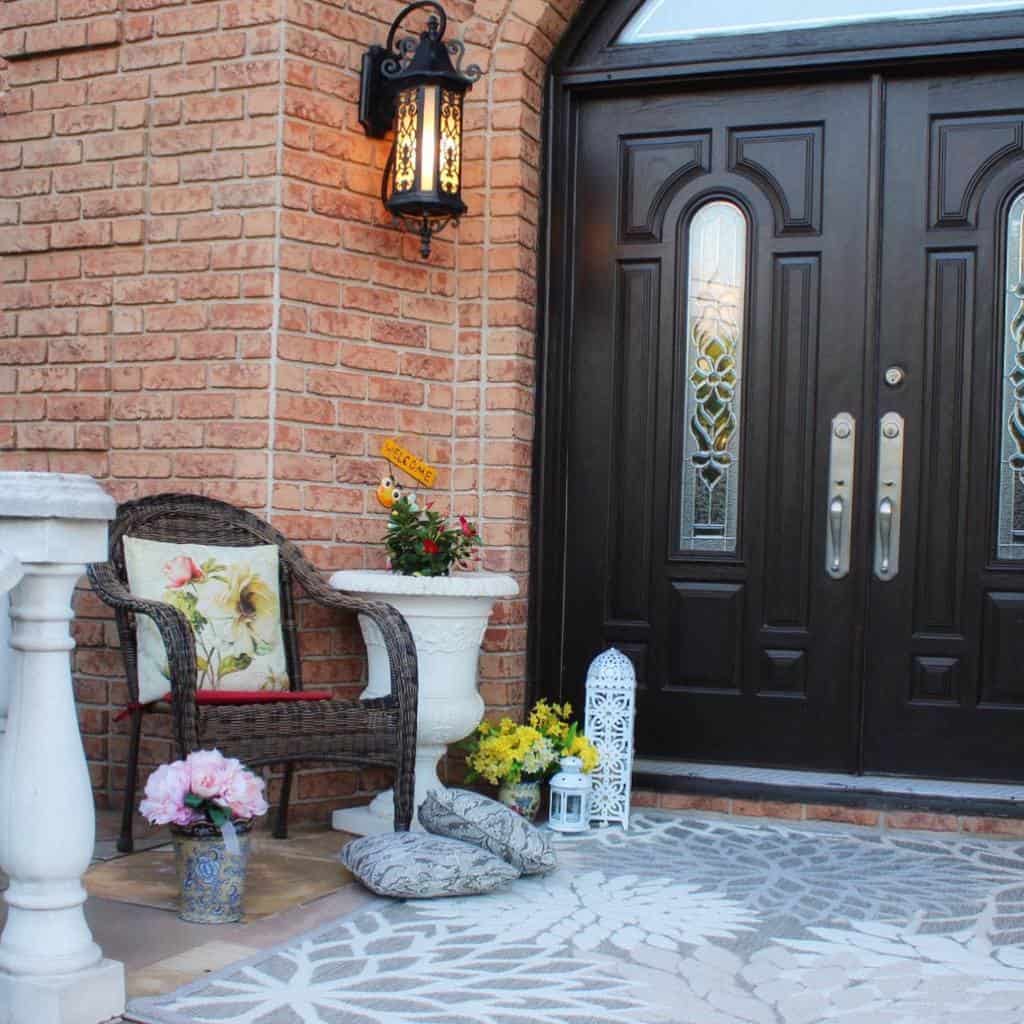 Cozy porch with a wicker chair, colorful cushions, a lantern, and flowers; double black doors and brick walls