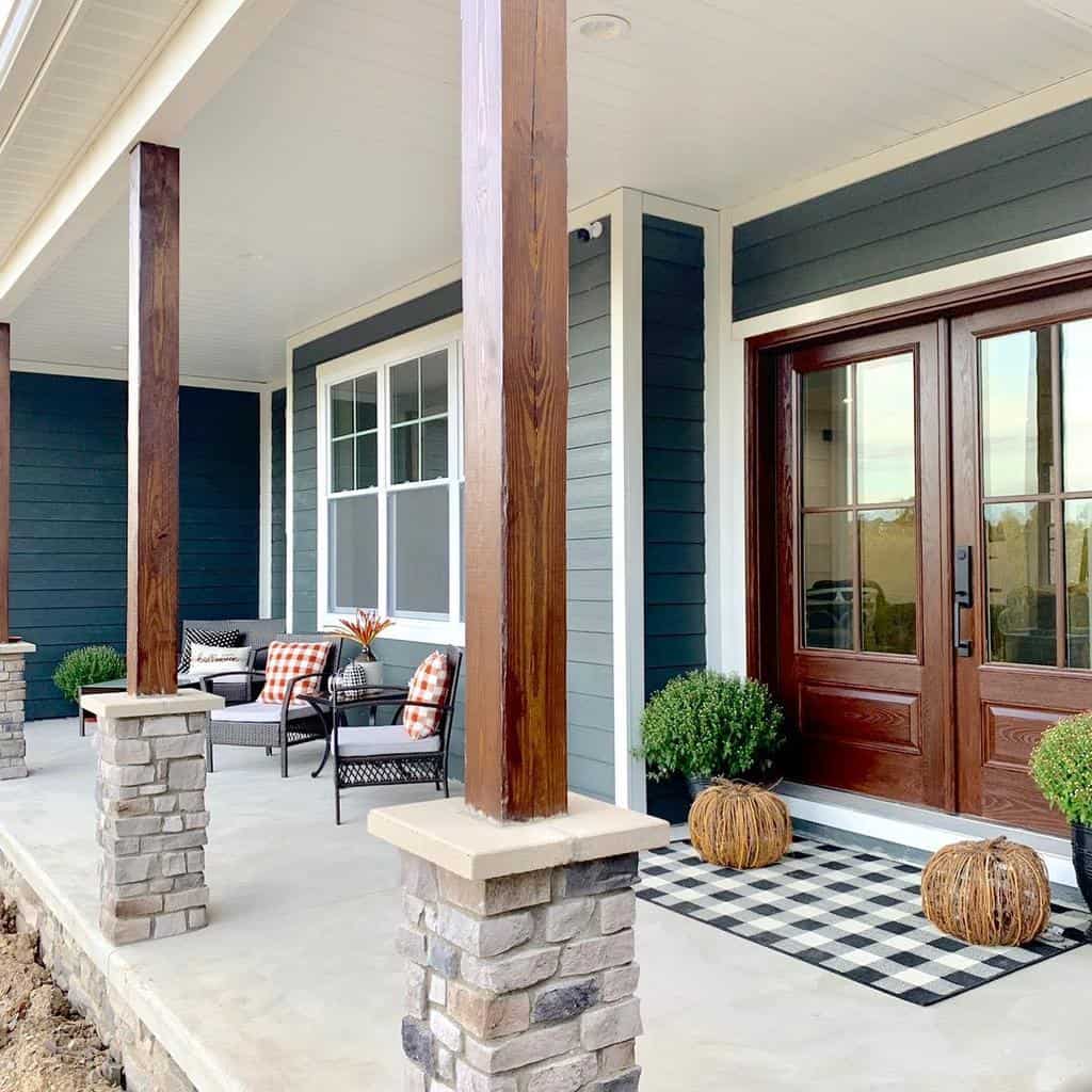 Modern house porch with wooden pillars, dark siding, double wooden doors, plaid doormat, potted plants, and checkered cushions on seating