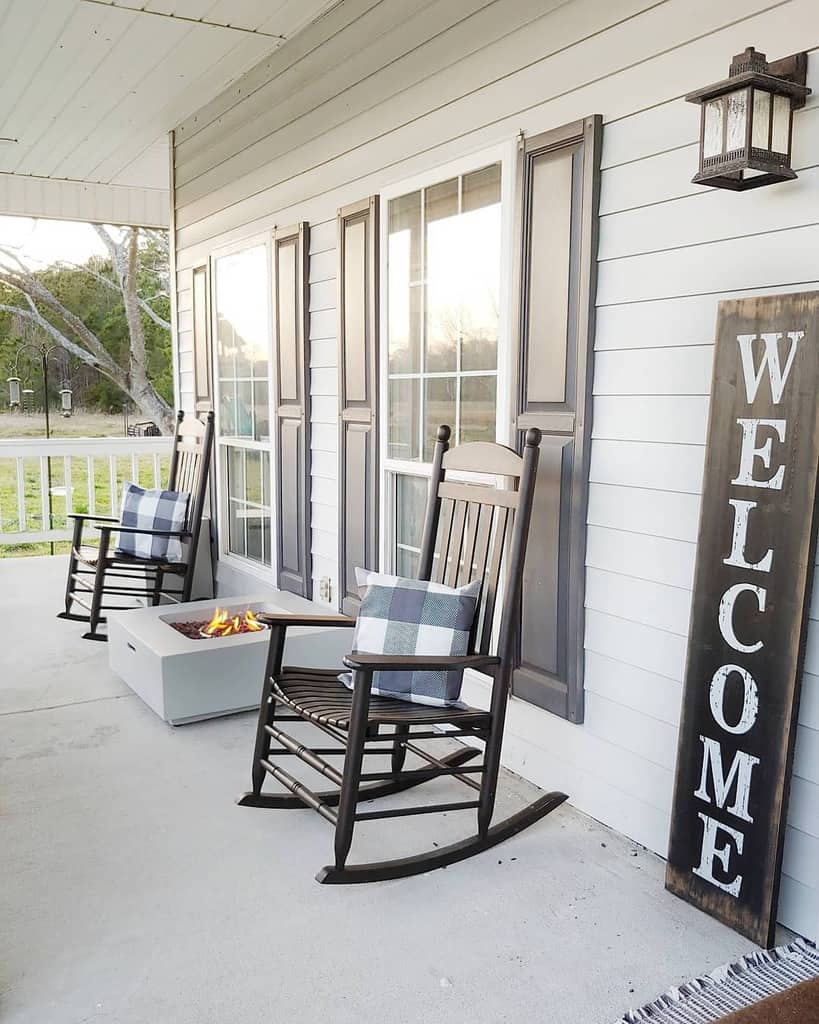 A cozy porch with two rocking chairs, a small firepit, plaid cushions, and a large "WELCOME" sign on a light-colored wall