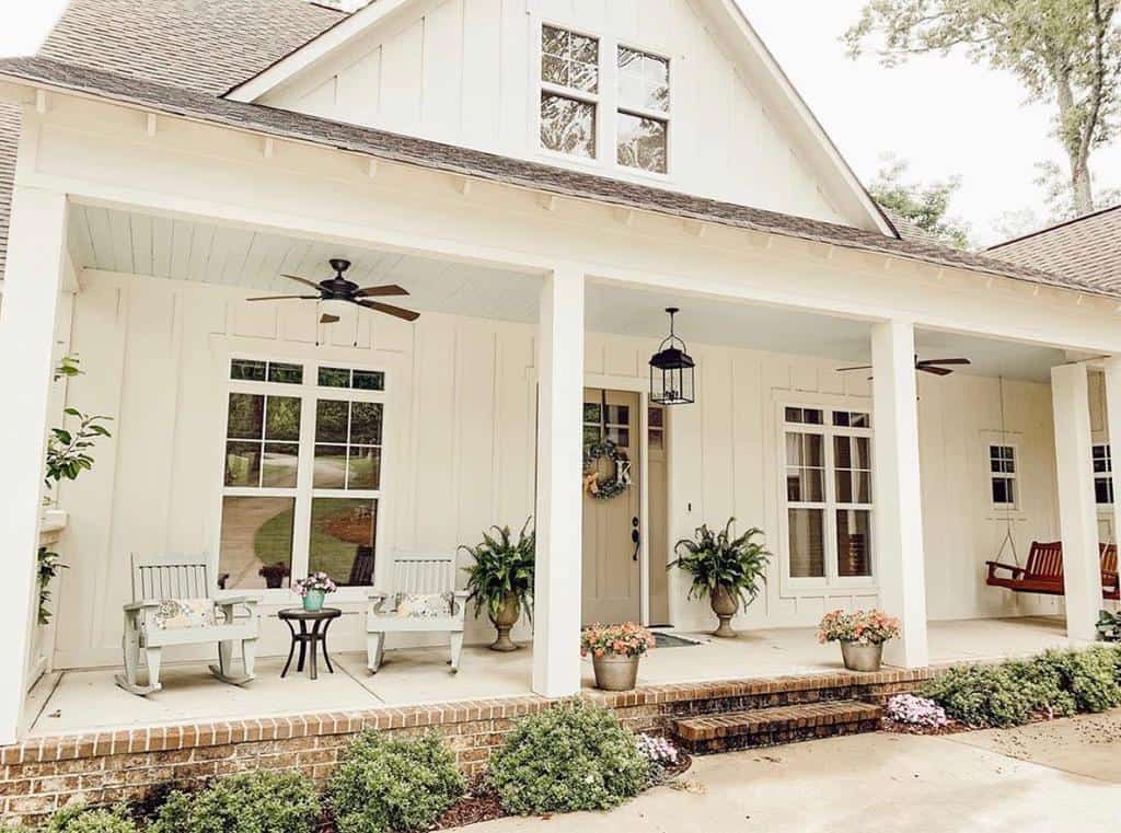 Front porch of a white house with potted plants, rocking chairs, a porch swing, and ceiling fans; steps lead up to the entrance