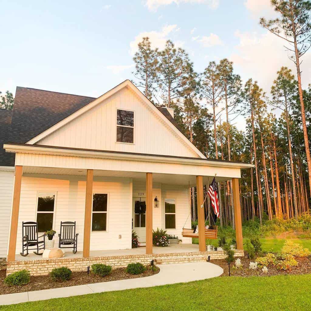 Charming farmhouse porch with wooden columns, rocking chairs, a swing, and an American flag, set against a peaceful wooded backdrop