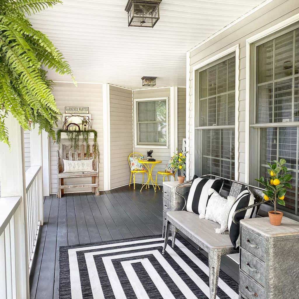 Charming porch with striped rug, metal bench, potted plants, and a small yellow table set. Hanging fern decorates the cozy space