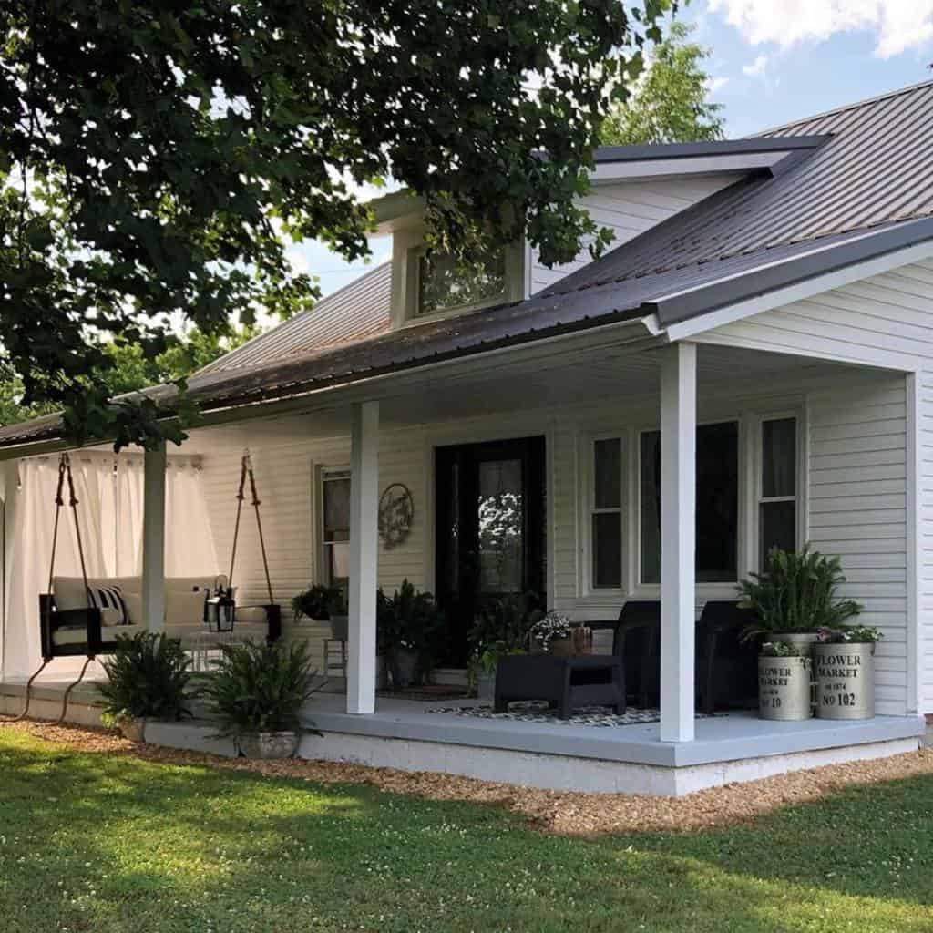 Charming farmhouse porch with a cozy swing, black front door, lush greenery, and inviting seating for relaxing outdoors