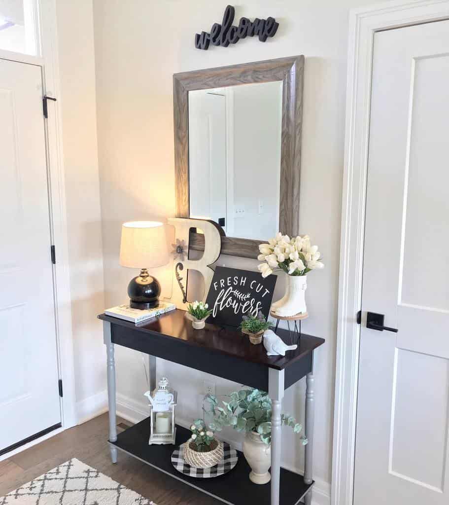 Farmhouse entryway with a black and gray console table, wood-framed mirror, warm lighting, floral decor, and a welcoming sign