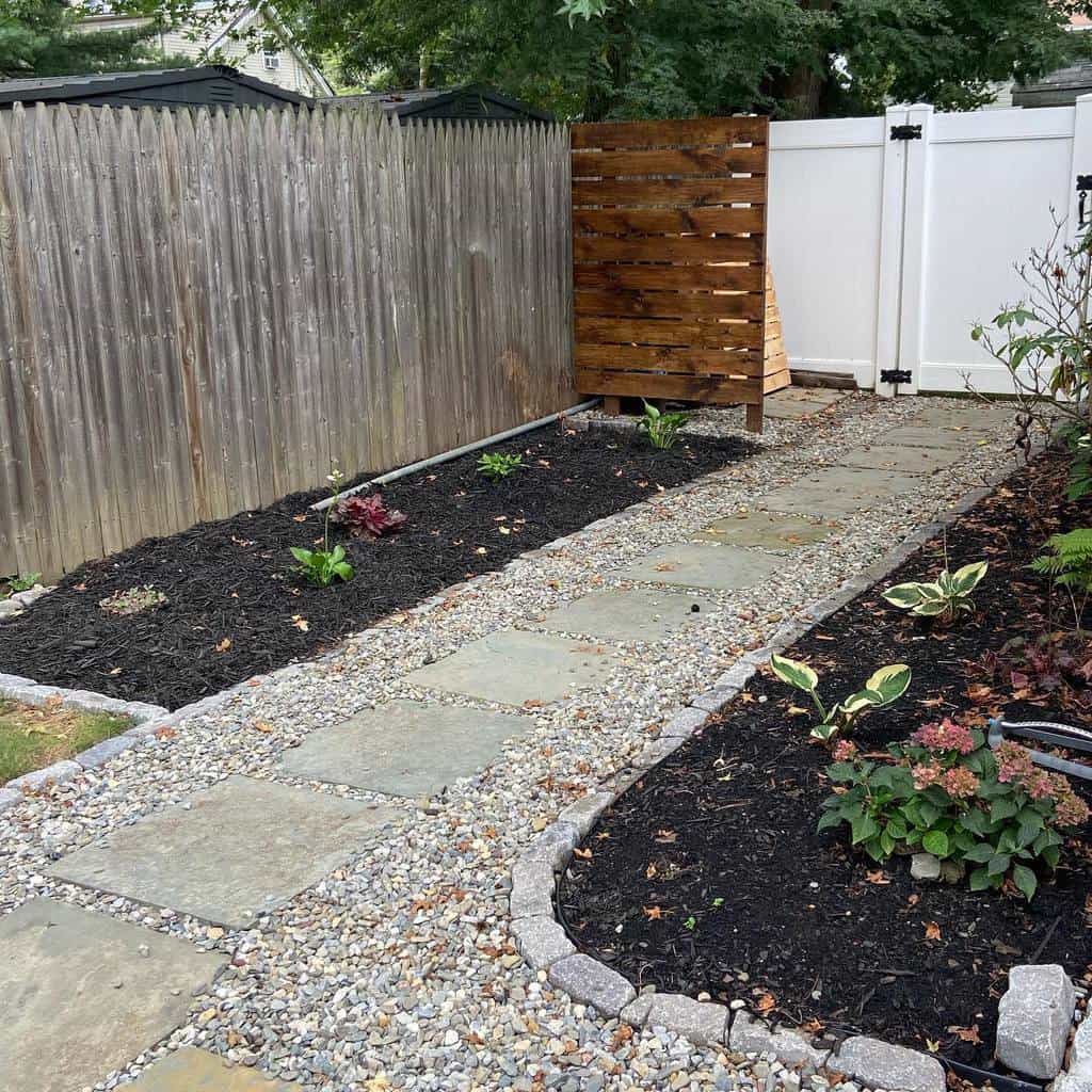 Garden path with large stone slabs and gravel, bordered by black mulch flower beds and a wooden and white fence