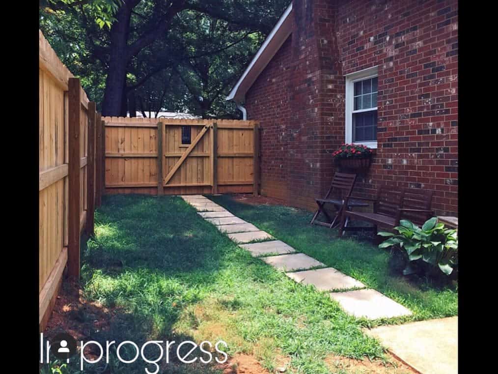 Side yard with a wooden fence, brick house, stepping stone path, green grass, outdoor seating, and a planter with flowers under the window