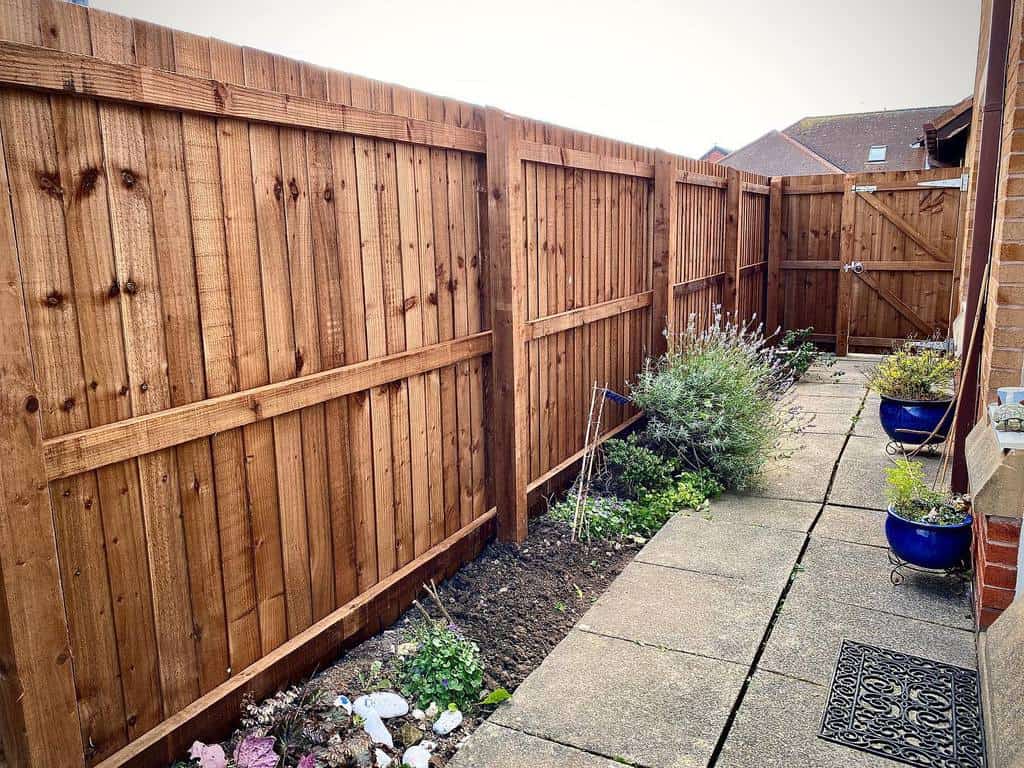 A narrow garden path with potted plants and wooden fencing on both sides leads to a closed gate