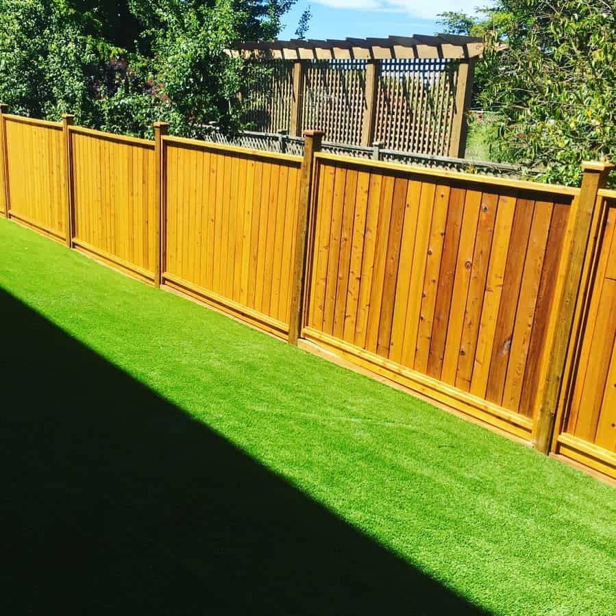 A neatly maintained yard with bright green artificial grass and a wooden fence, featuring trees and a pergola in the background