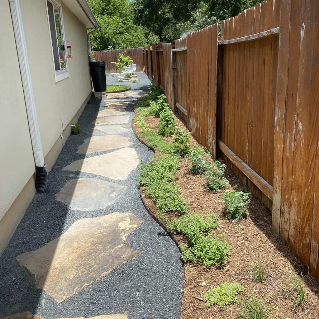 Stone pathway beside a house with a wooden fence on the right and green plants along the edge of the path