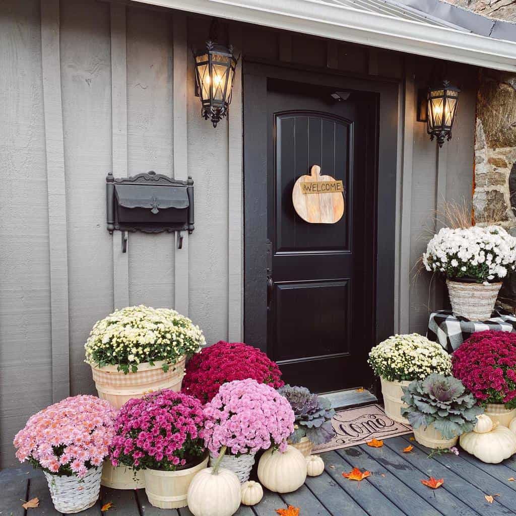 Front porch with a black door, welcome sign, flower pots, pumpkins, and lanterns; autumn-themed decor and charming vibe