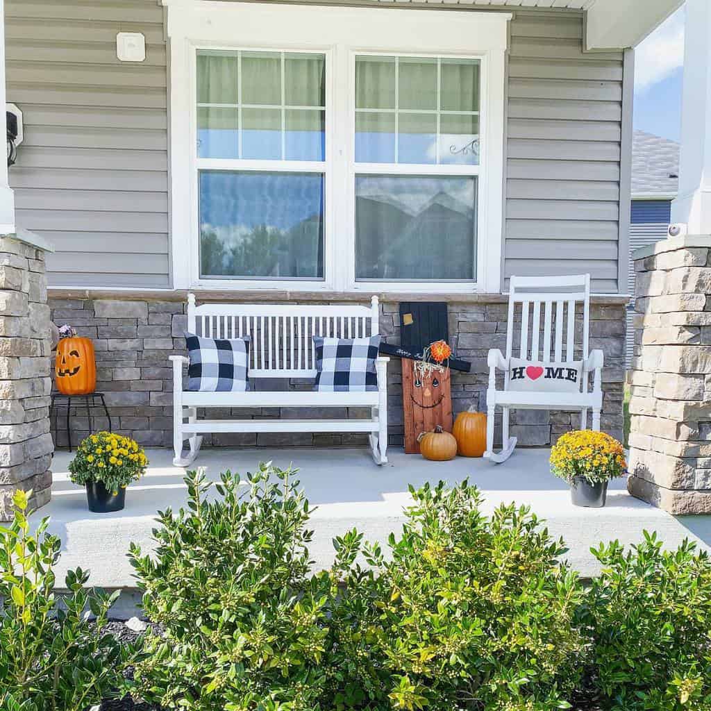 A cozy residential porch with a white swing, rocking chair, pumpkins, and potted flowers, featuring a Jack-o'-lantern and checkered pillows for added charm