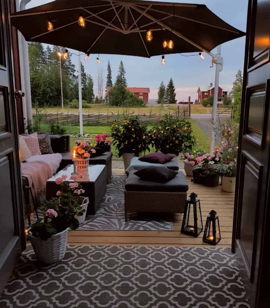 Front porch with wicker seating, cushions, lanterns, flowers, and a large umbrella overlooking a garden and countryside view at dusk