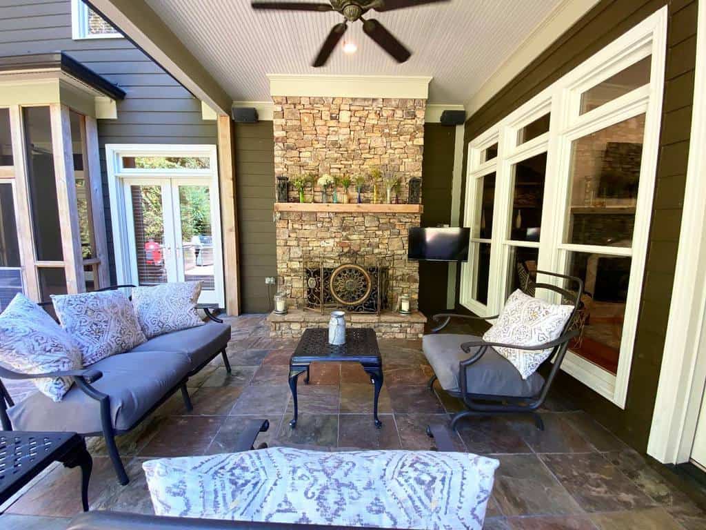 Porch with stone fireplace, ceiling fan, cushioned seating, and a small TV mounted on the wall, surrounded by large windows