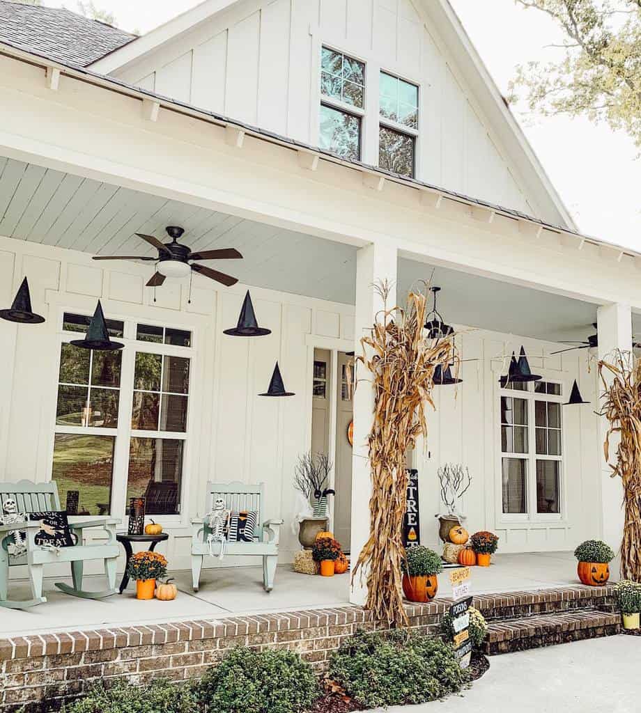 Front porch with rocking chairs, pumpkins, hanging witch hats, and dried corn stalks; white house decor for Halloween