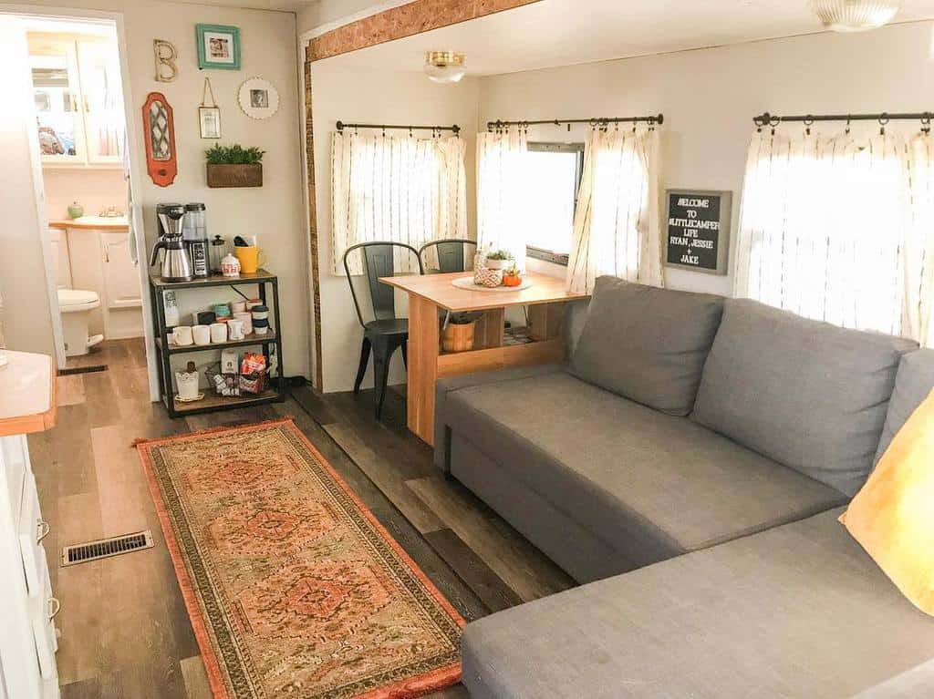 Cozy small living space with gray sofa, dining area, coffee station, and decorative rug. Bright curtains allow natural light inside.