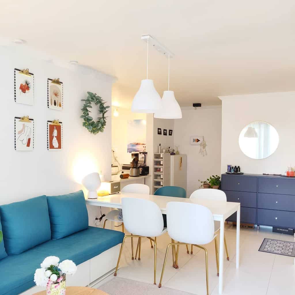 Bright living room with a teal sofa, white dining table, modern chairs, wall art, greenery wreath, and pendant lights.