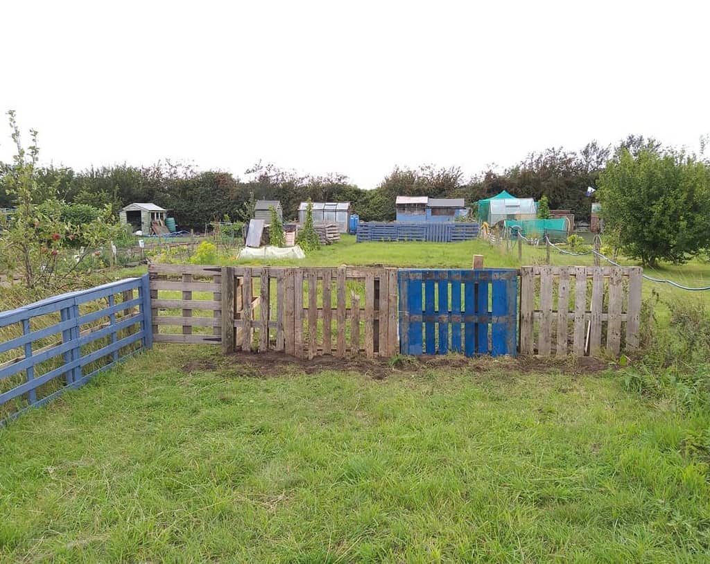 Rustic DIY pallet fence with a mix of natural and blue-painted wood, enclosing a small garden plot in a lush rural setting