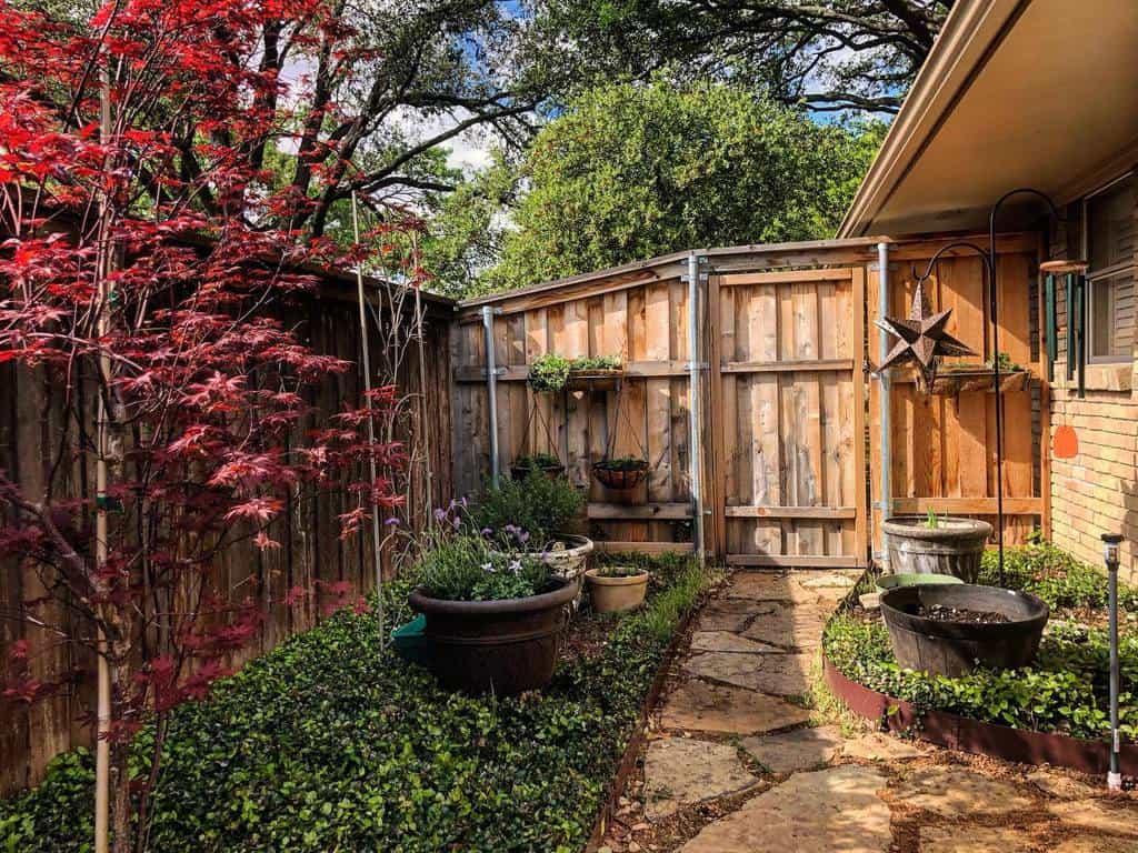 Cozy side yard with a stone pathway, potted plants, lush greenery, a red maple tree, and a rustic wooden fence with hanging decor