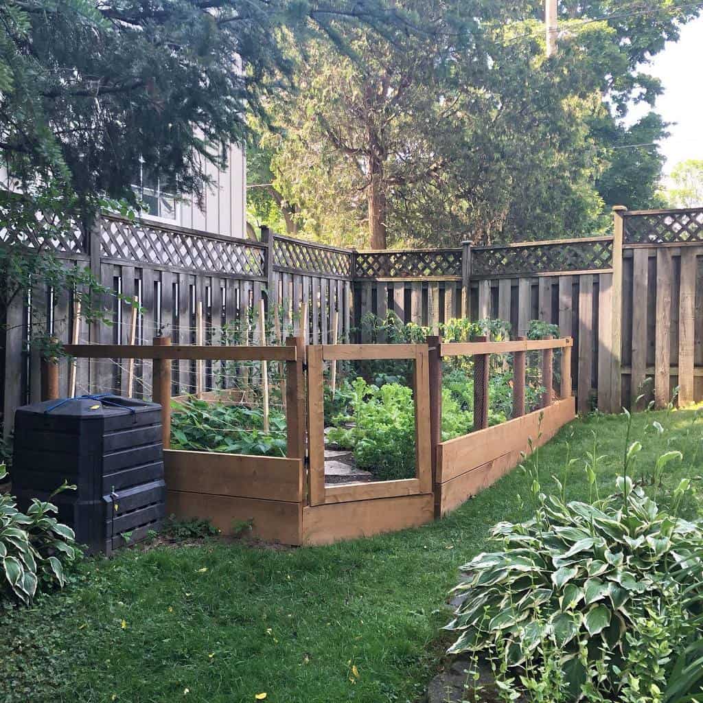 A small backyard garden with raised wooden beds, surrounded by a wooden fence, lush greenery, and a black compost bin