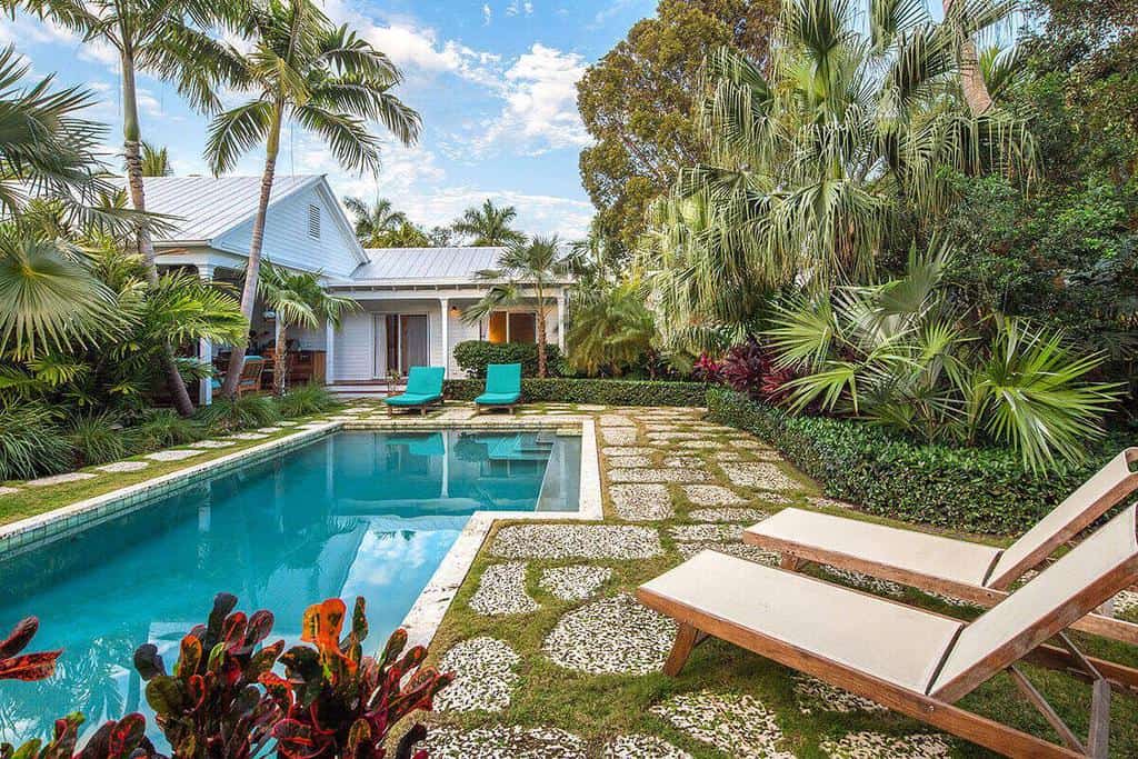 Tropical backyard with a pool, two turquoise lounge chairs, lush greenery, and a white house in the background