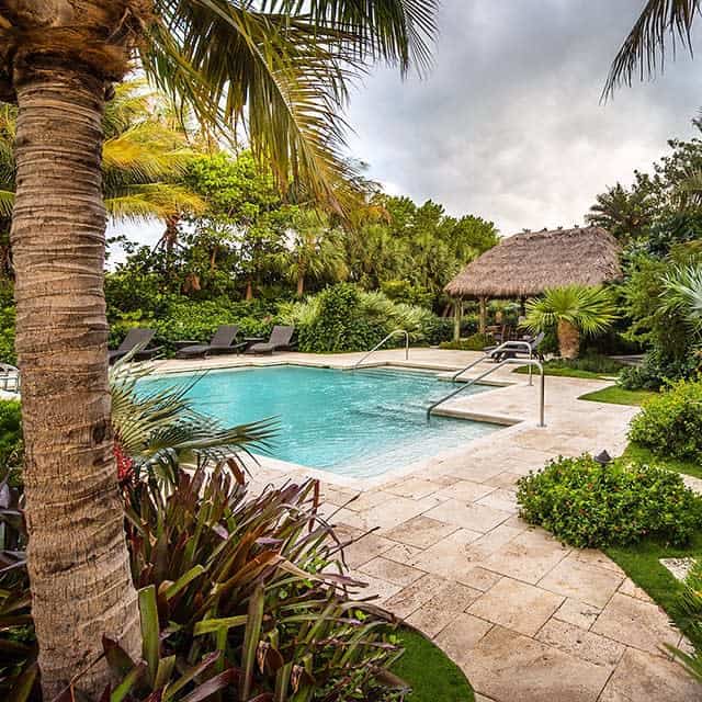 A tropical pool scene with a thatched hut, palm trees, and lounge chairs surrounded by lush greenery under a cloudy sky