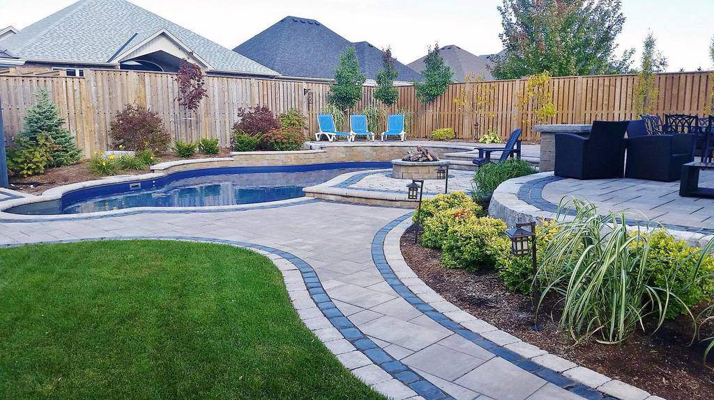 Backyard with a curved pool, patio, lawn, and lounge chairs surrounded by a wooden fence and diverse plants